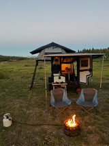 Twilight RV rental camping scene: a camper trailer with a rooftop tent, chairs, and a propane fire pit.