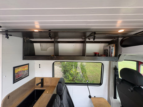 Cozy rv rental camper van interior: wood-slat ceiling, dark overhead storage with books, kitchenette, and a scenic window view.