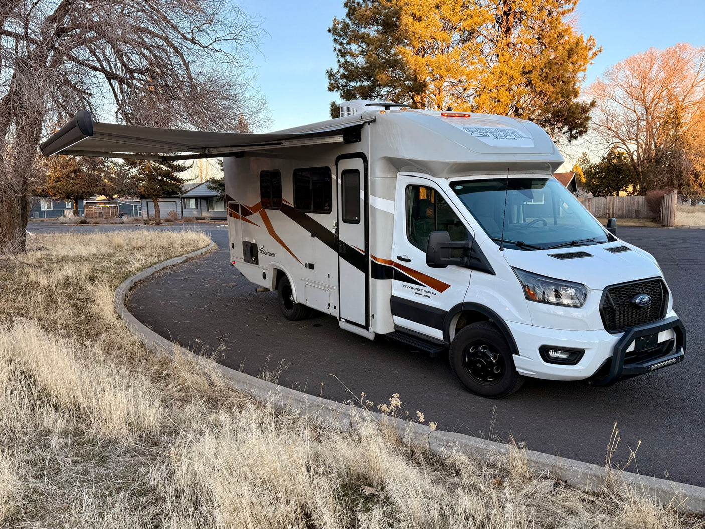 A white Coachmen RV rental on Ford Transit chassis with awning extended, parked by dry grass.