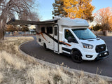 A white Coachmen RV rental on Ford Transit chassis with awning extended, parked by dry grass.