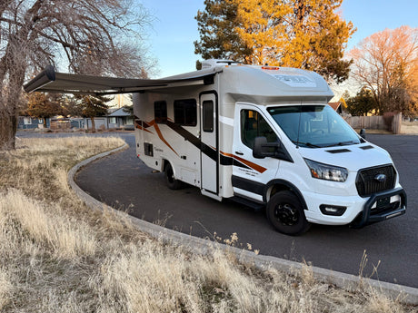 A white Coachmen RV rental on Ford Transit chassis with awning extended, parked by dry grass.