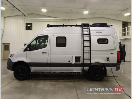 Silver Mercedes-Benz Sprinter rv rental camper van with roof rack, ladder, and off-road tires, charging indoors.