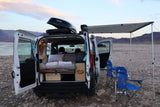 Enjoy lakeside camping! This RV rental camper van is open, revealing a bed & storage. An awning and chairs are set up on the rocky shore.
