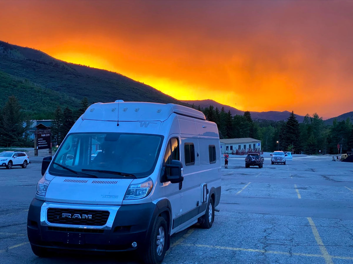 Vibrant orange sunset illuminates mountains behind a sleek grey RV rental parked in Park City. Other vehicles are in the lot.|Experience nature with this silver pop-top camper van **rv rental** parked by a serene lake and green trees under a blue sky.|Modern RV rental interior: integrated fridge