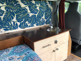 Custom **RV rental** interior: wooden kitchenette with sink, a cozy bed, and vibrant tropical leaf curtains.