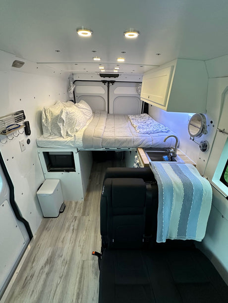 Inside an RV rental: a modern camper van with a raised bed, kitchenette sink, overhead storage, and light wood flooring.
