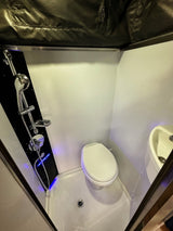 A bright, compact RV rental bathroom showing a shower panel with blue accent lighting, a toilet, and a small sink.