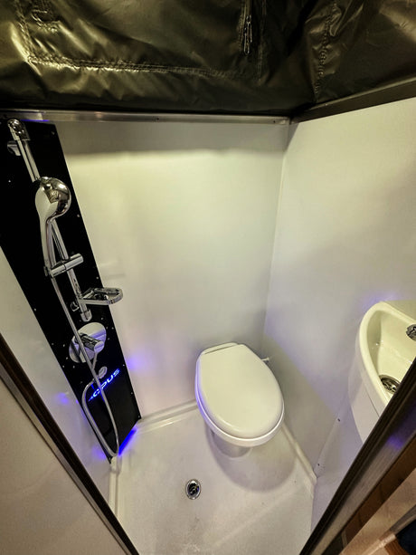 A bright, compact RV rental bathroom showing a shower panel with blue accent lighting, a toilet, and a small sink.