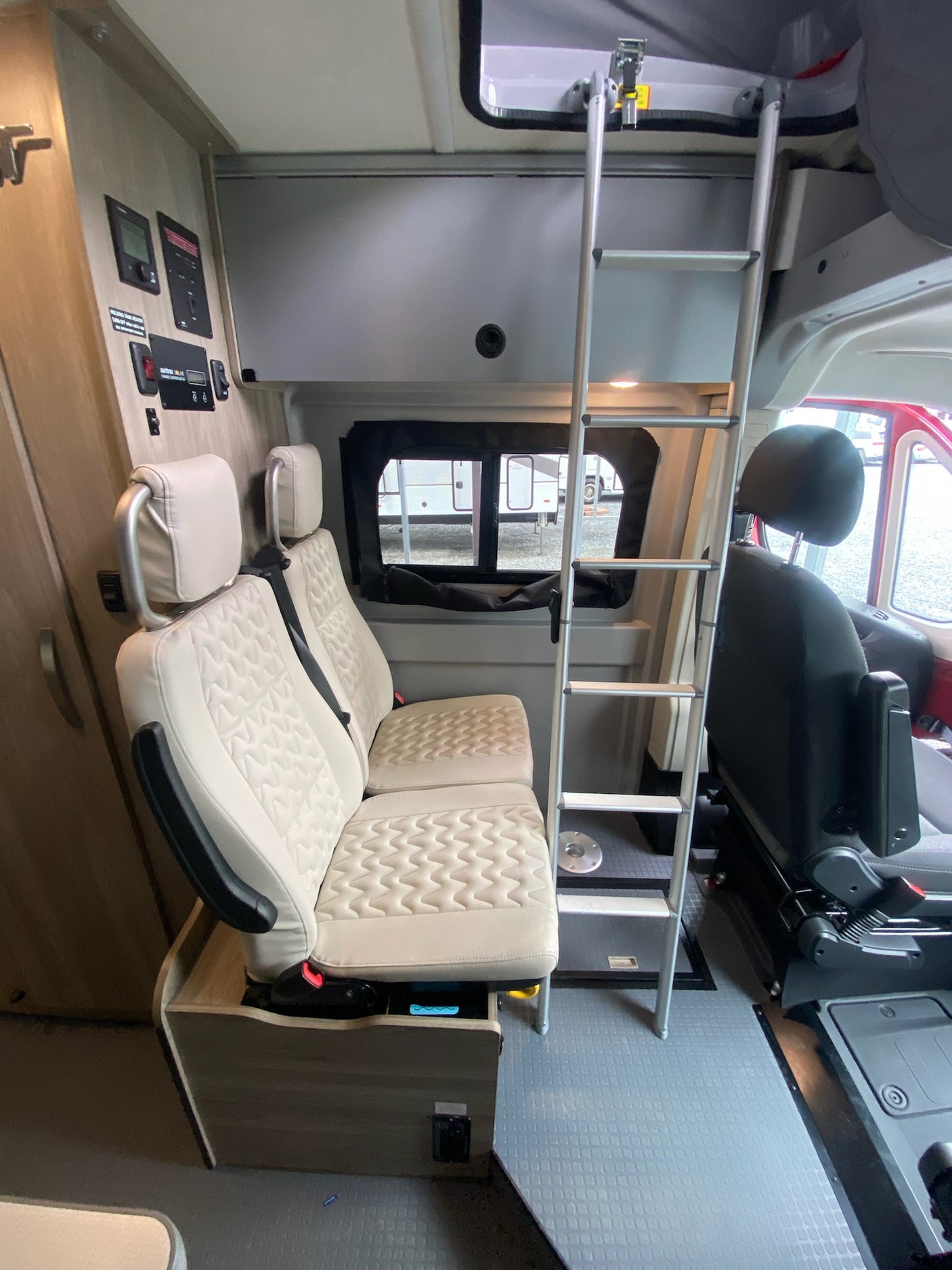 Interior view of an RV rental showing beige passenger seats, ladder to an overhead bunk, and wall-mounted controls.