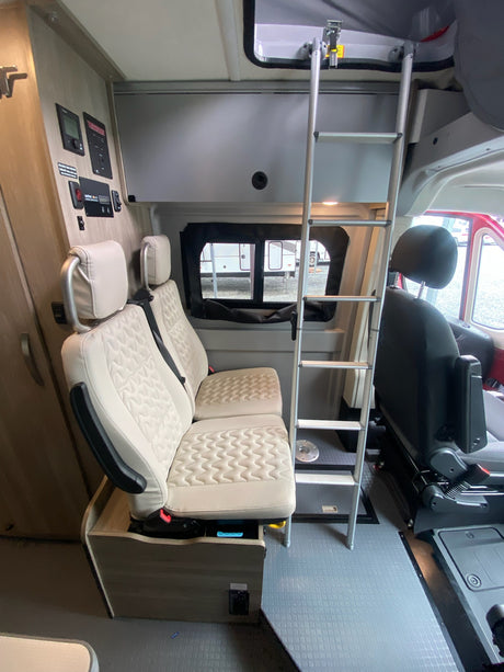 Interior view of an RV rental showing beige passenger seats, ladder to an overhead bunk, and wall-mounted controls.