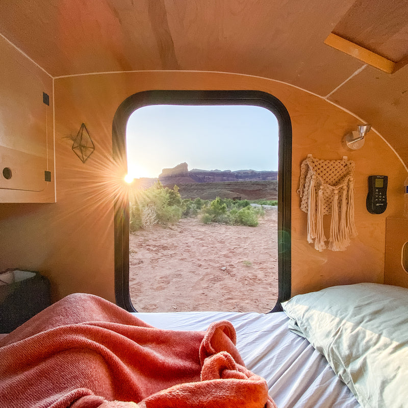 Warm light streams into an RV rental from a desert sunset, illuminating a cozy bed. Mountains visible through the window. — photo 18