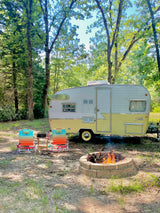 A cozy RV rental campsite with a yellow and white vintage camper, outdoor chairs, and a warm fire pit for glamping.