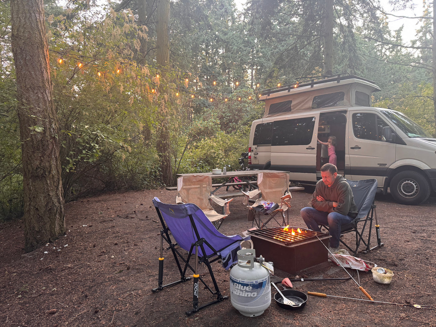 Family enjoying an RV rental campsite. Pop-top camper van, string lights, campfire cooking, and cozy camping chairs.