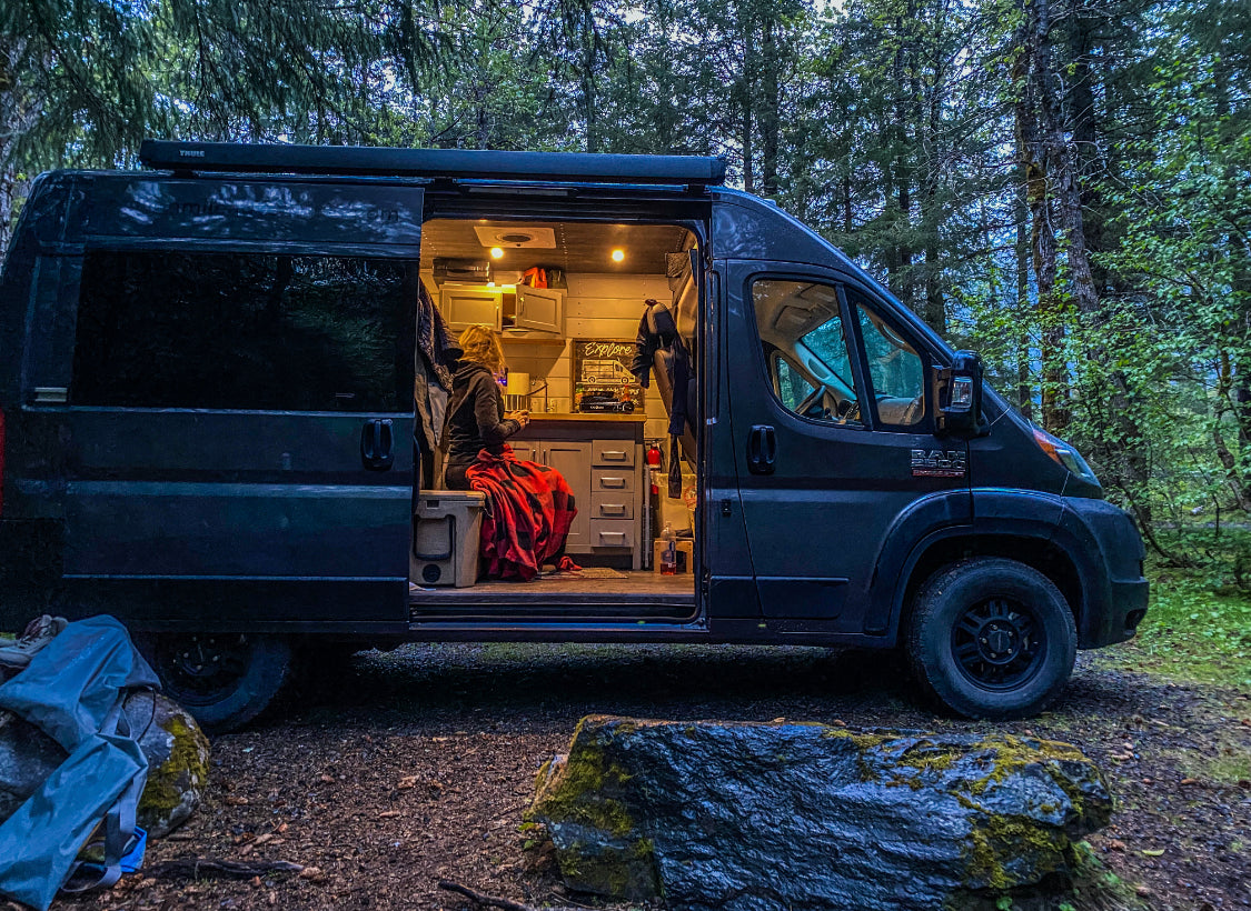 An RV rental camper van parked in a forest, its side open to reveal a cozy, lit interior with a person wrapped in a red blanket.