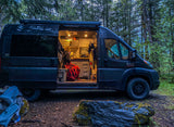 An RV rental camper van parked in a forest, its side open to reveal a cozy, lit interior with a person wrapped in a red blanket.