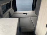 Interior of an rv rental features a gray convertible bed/seating area, a white counter, and window with a dark shade.