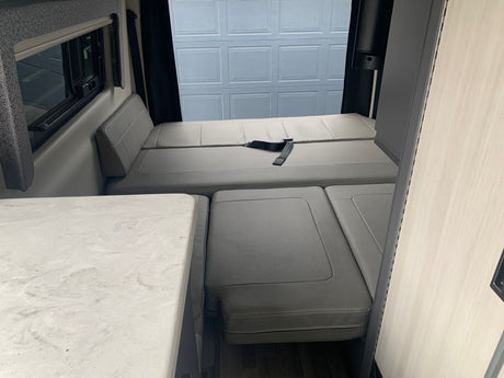 Interior of an rv rental features a gray convertible bed/seating area, a white counter, and window with a dark shade.