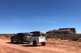 RV rental: A black Jeep tows an RV-style trailer with a rooftop tent on a red dirt road in an arid desert landscape.