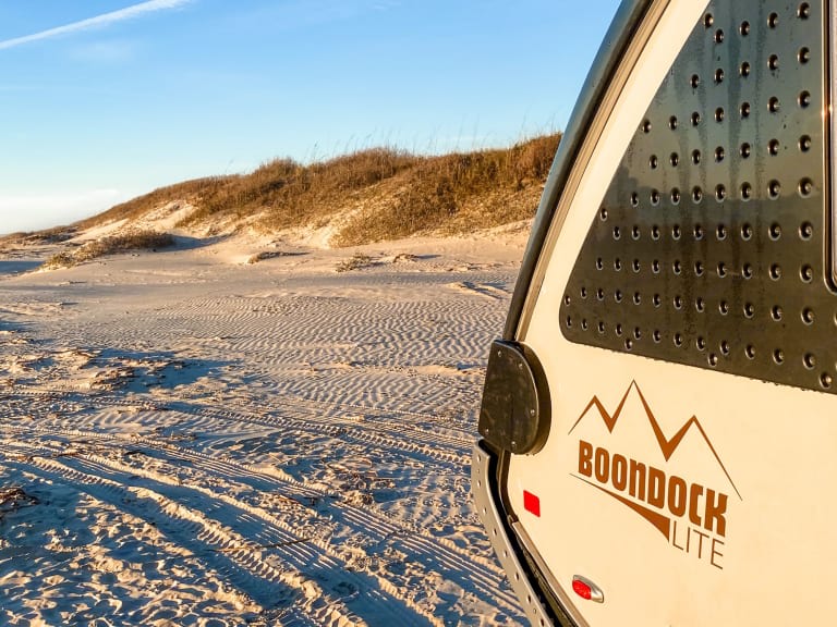 This RV rental, a Boondock Lite, on a sandy beach near dunes offers perfect outdoor camping and adventure.
