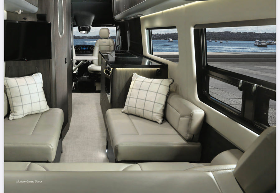 Experience luxury RV rental with this spacious, modern greige interior showing comfy seating, kitchen, and a scenic view of boats on the water.