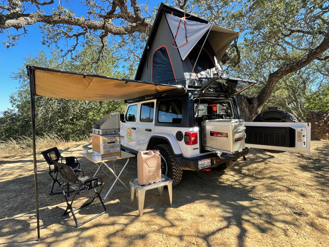 White Jeep RV rental fully equipped with rooftop tent, extended awning, outdoor kitchen, chairs, and fridge setup.