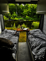 Cozy RV rental bedroom with twin beds, coffee, and a book, offering a stunning view of a serene river and lush forest.