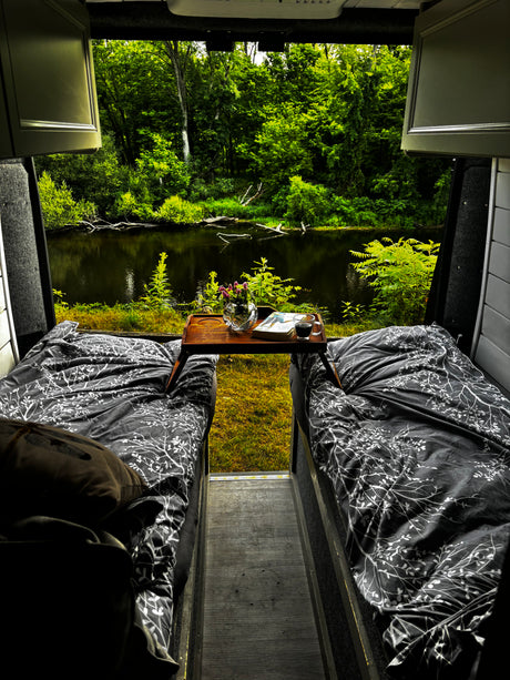 Cozy RV rental bedroom with twin beds, coffee, and a book, offering a stunning view of a serene river and lush forest.