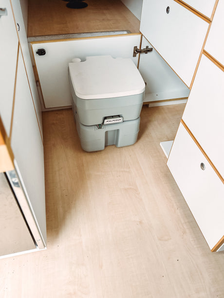 Portable ALPCour toilet on light wood flooring in an RV rental's neat, white cabinet interior.