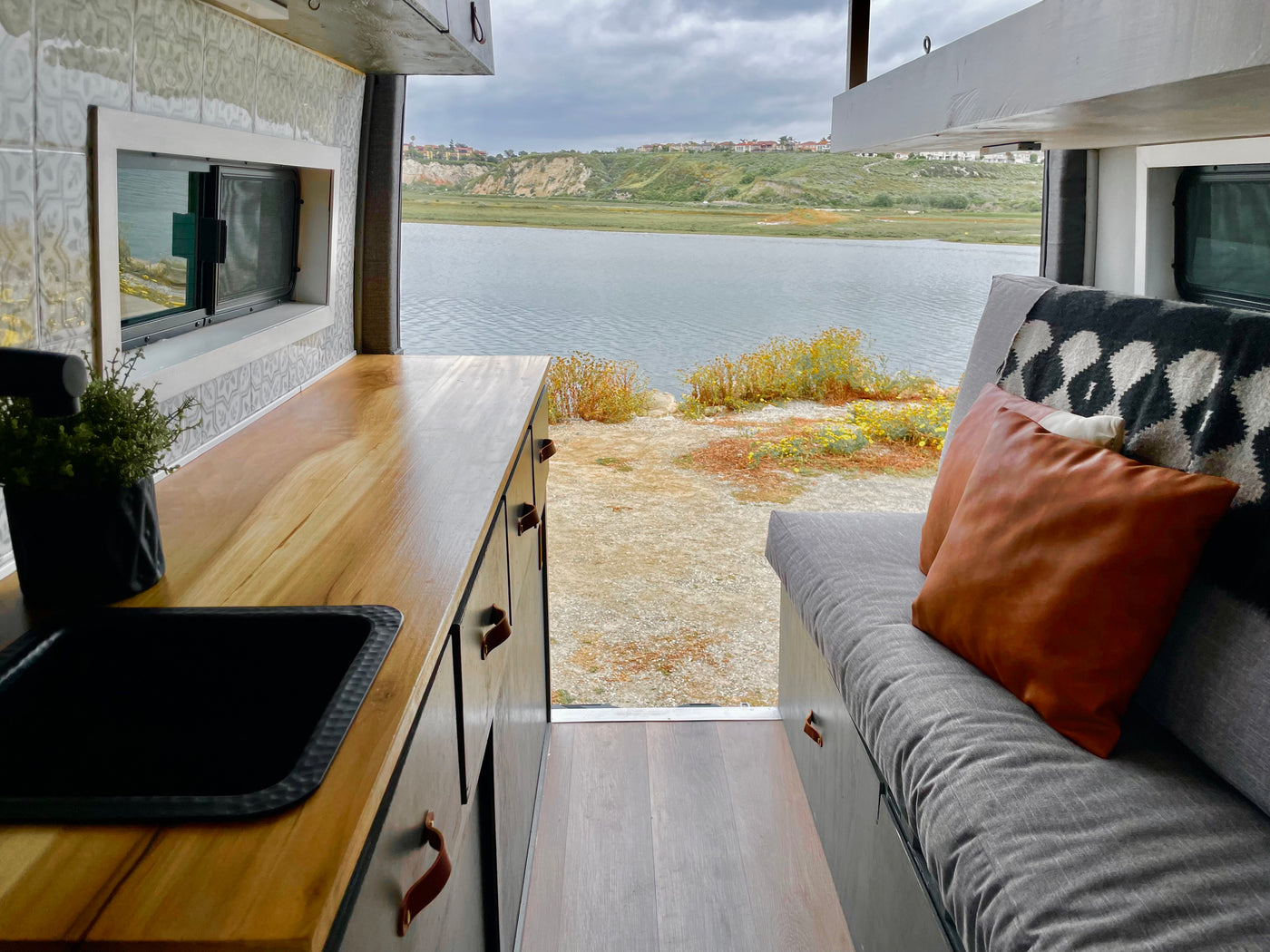 This RV rental features a comfortable interior with a kitchenette and couch, offering serene views of a calm bay and distant hills. — photo 10