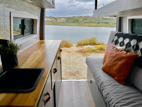 This RV rental features a comfortable interior with a kitchenette and couch, offering serene views of a calm bay and distant hills.