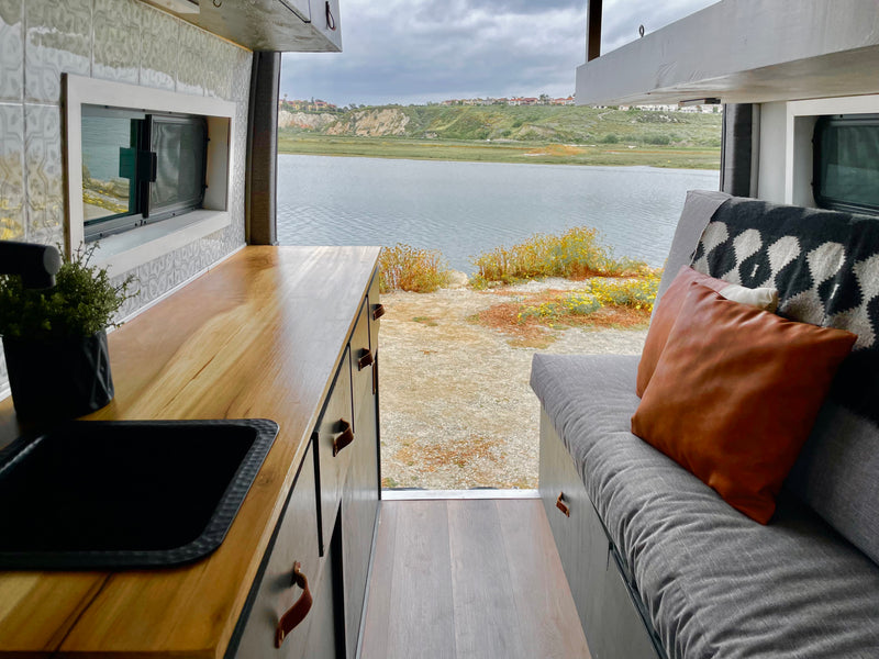 This RV rental features a comfortable interior with a kitchenette and couch, offering serene views of a calm bay and distant hills. — photo 10