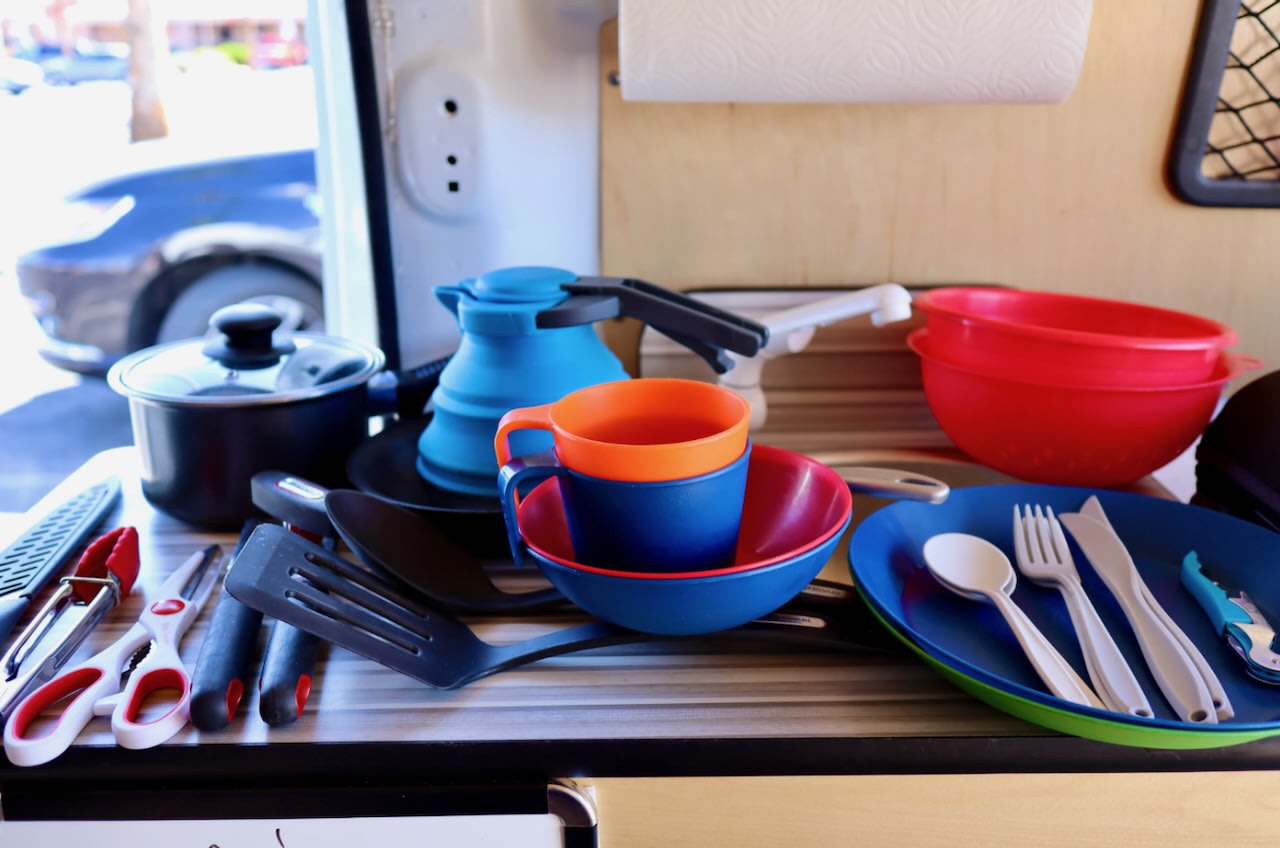 A compact **RV rental** kitchen featuring a pot, collapsible kettle, bright plastic dishes, and various **camping kitchen essentials**.