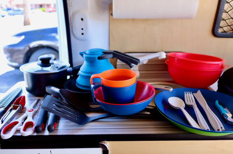 A compact **RV rental** kitchen featuring a pot, collapsible kettle, bright plastic dishes, and various **camping kitchen essentials**.