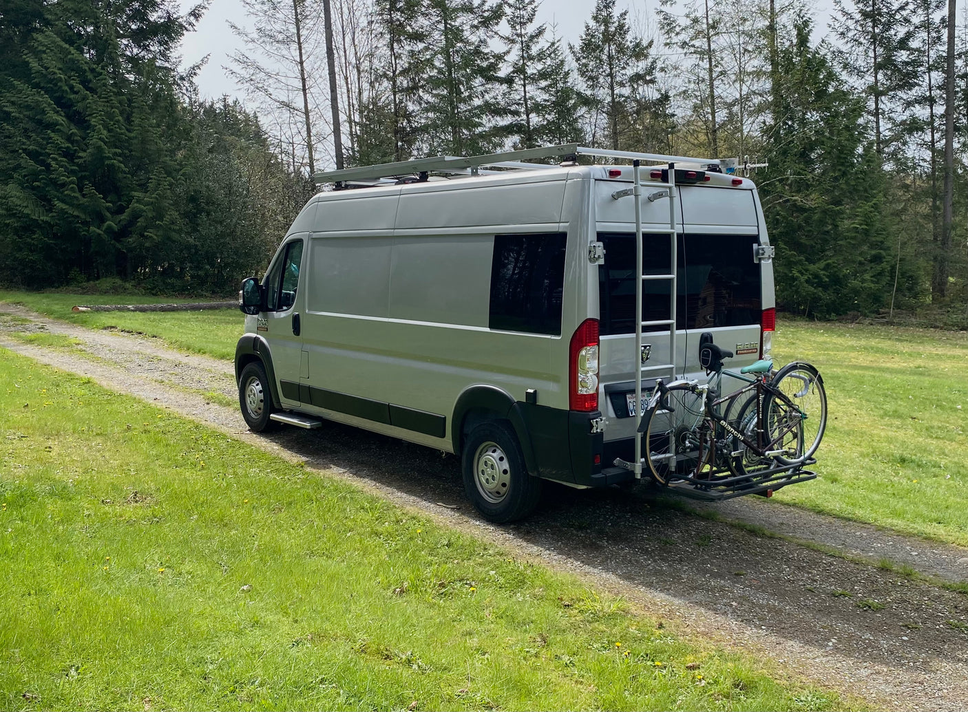 RV rental camper van with roof rack and bikes on a path. Tall trees & green grass for outdoor adventure.