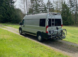 RV rental camper van with roof rack and bikes on a path. Tall trees & green grass for outdoor adventure.
