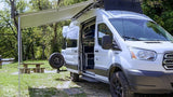 White rv rental camper van with extended awning at a wooded campsite.
