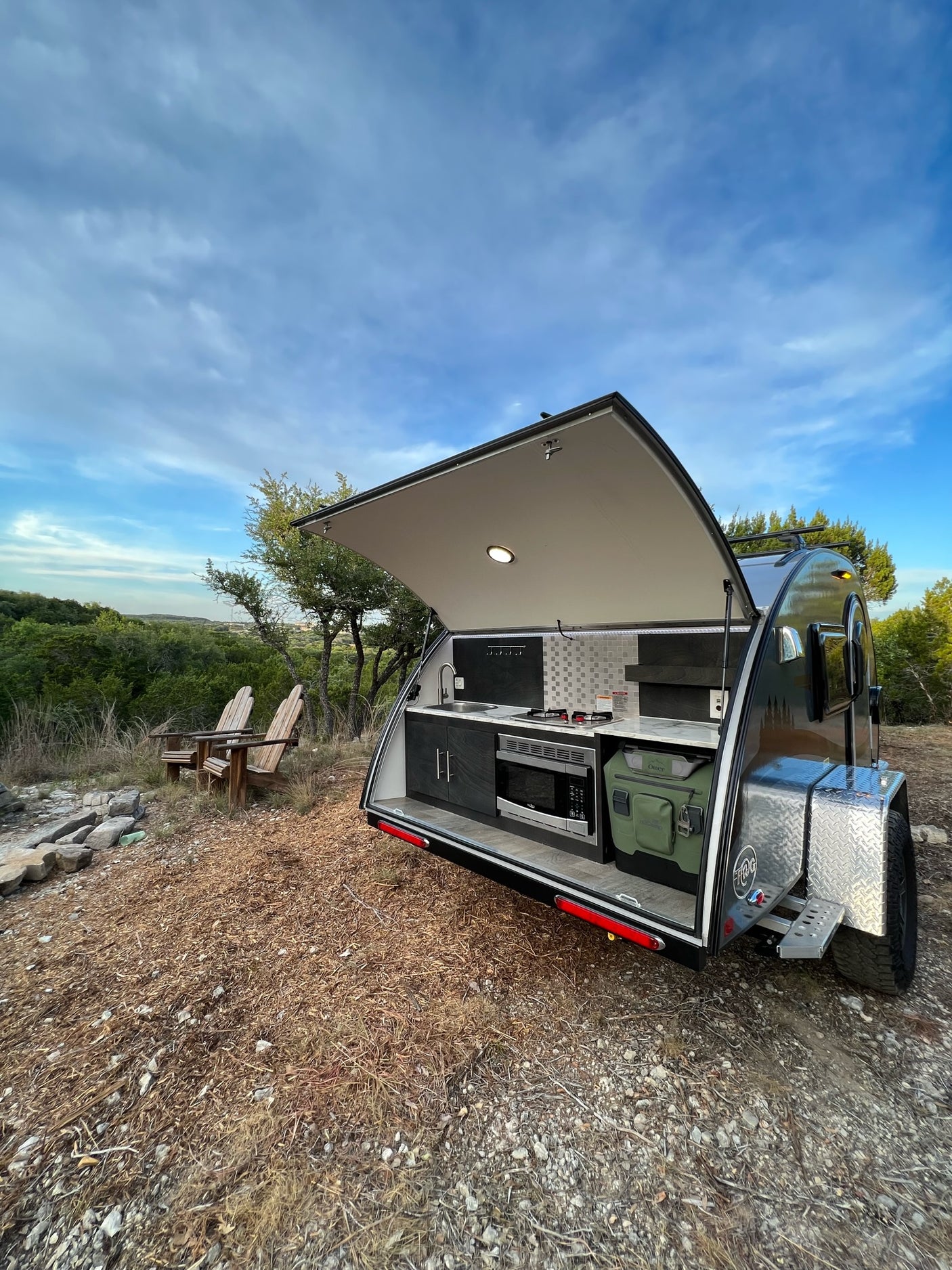 A tear-drop RV rental's open rear kitchen reveals a stove, sink, and microwave. Two chairs face a scenic view.