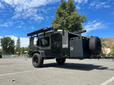 This dark grey Hiker Trailer, an ideal RV rental for outdoor adventures, features an awning, propane tank, and spare tire.