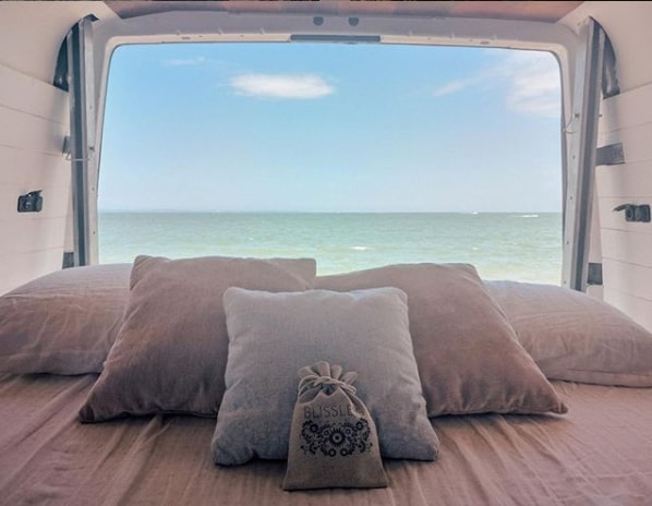 Cozy RV rental bed with pillows faces the ocean, offering tranquil views. Relax with your Blissle pouch.