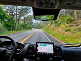 Inside an RV rental, driving on a scenic, winding road. Dashboard GPS visible, a red car reflected in the rearview mirror.