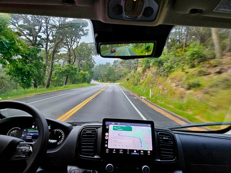 Inside an RV rental, driving on a scenic, winding road. Dashboard GPS visible, a red car reflected in the rearview mirror.