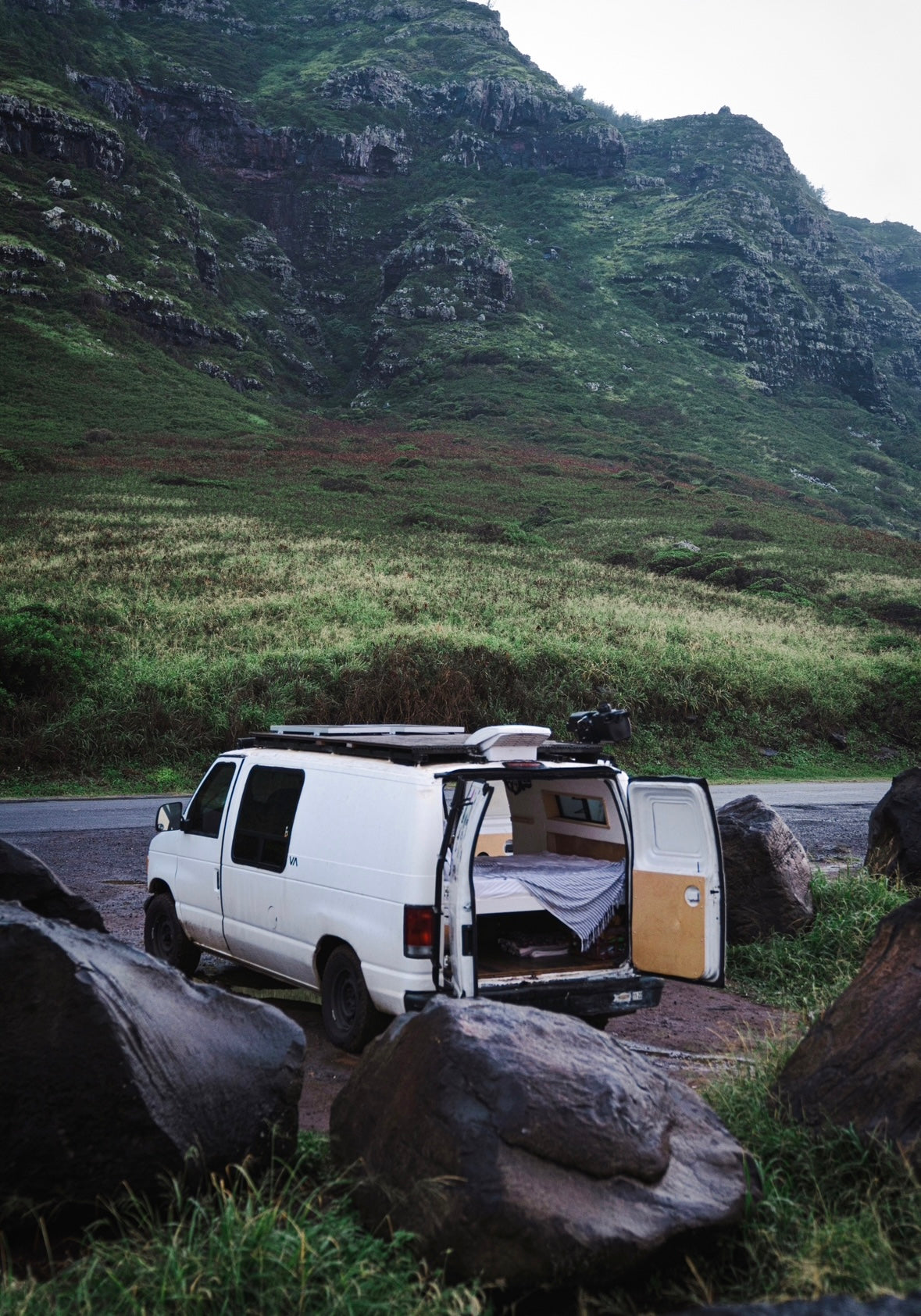 White camper van with open back, bed visible, parked among rocks with a vast green mountain behind. Perfect RV rental for nature travel.
