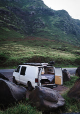 White camper van with open back, bed visible, parked among rocks with a vast green mountain behind. Perfect RV rental for nature travel.