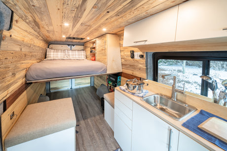 Inside a modern RV rental: a cozy wooden interior with a raised bed, functional kitchen, and bench seating for travel.