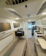 Interior of a modern rv rental, showing kitchen, dining area, couch, and TV, with light wood floors and overhead storage.