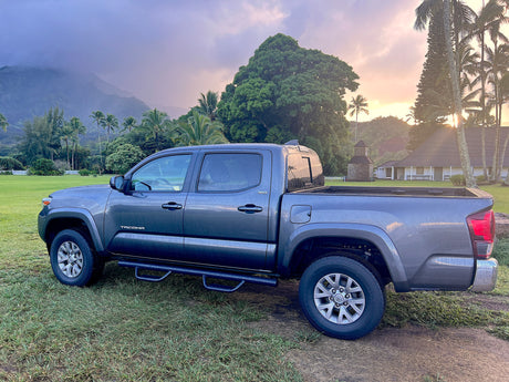 A dark grey Toyota Tacoma pickup sits on grassy grounds, framed by tropical mountains and a sunset glow. Ideal for local adventures, or try an rv rental.