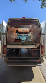 This RV rental showcases a bright, wood-paneled camper van interior with a comfortable bed, under-bed storage, and front cab view.