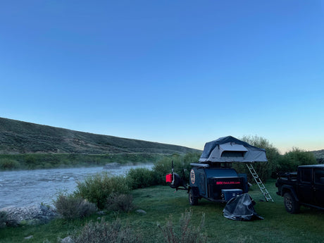An RV rental trailer with a rooftop tent and truck by a misty river and green hills.