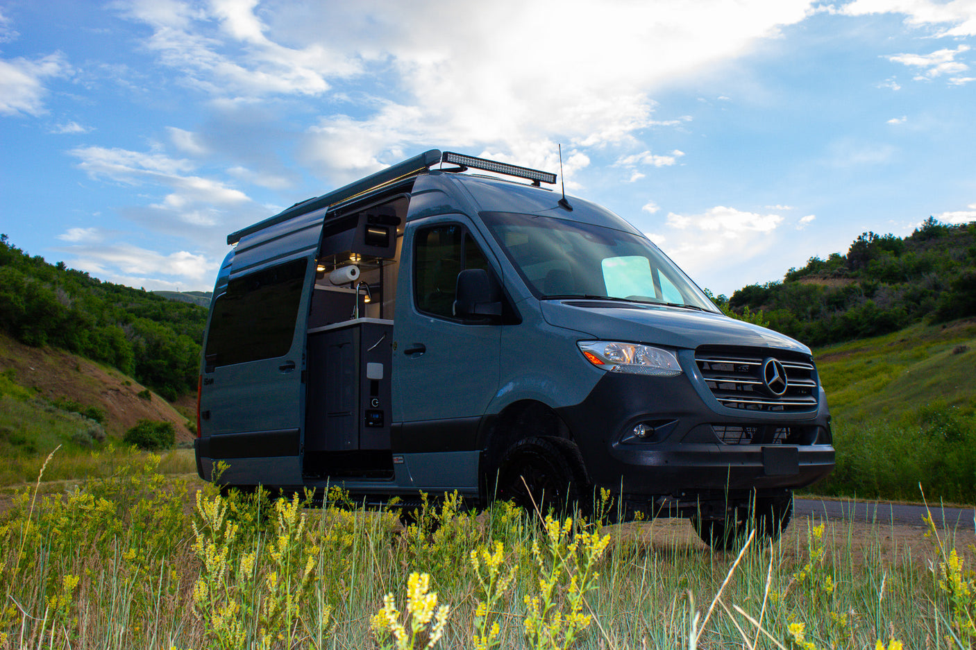 Explore the outdoors! Modern gray Sprinter camper van rv rental with open side door showing kitchenette, parked in grassy hills.