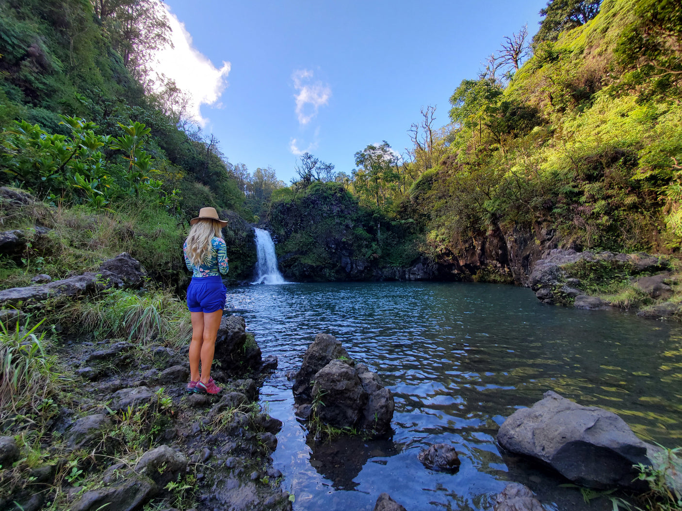Woman on rocks by a vibrant blue pool and waterfall amidst lush greenery. Perfect RV rental destination!
